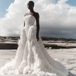 Woman in a white wedding dress with feathered details standing on a rocky beach.