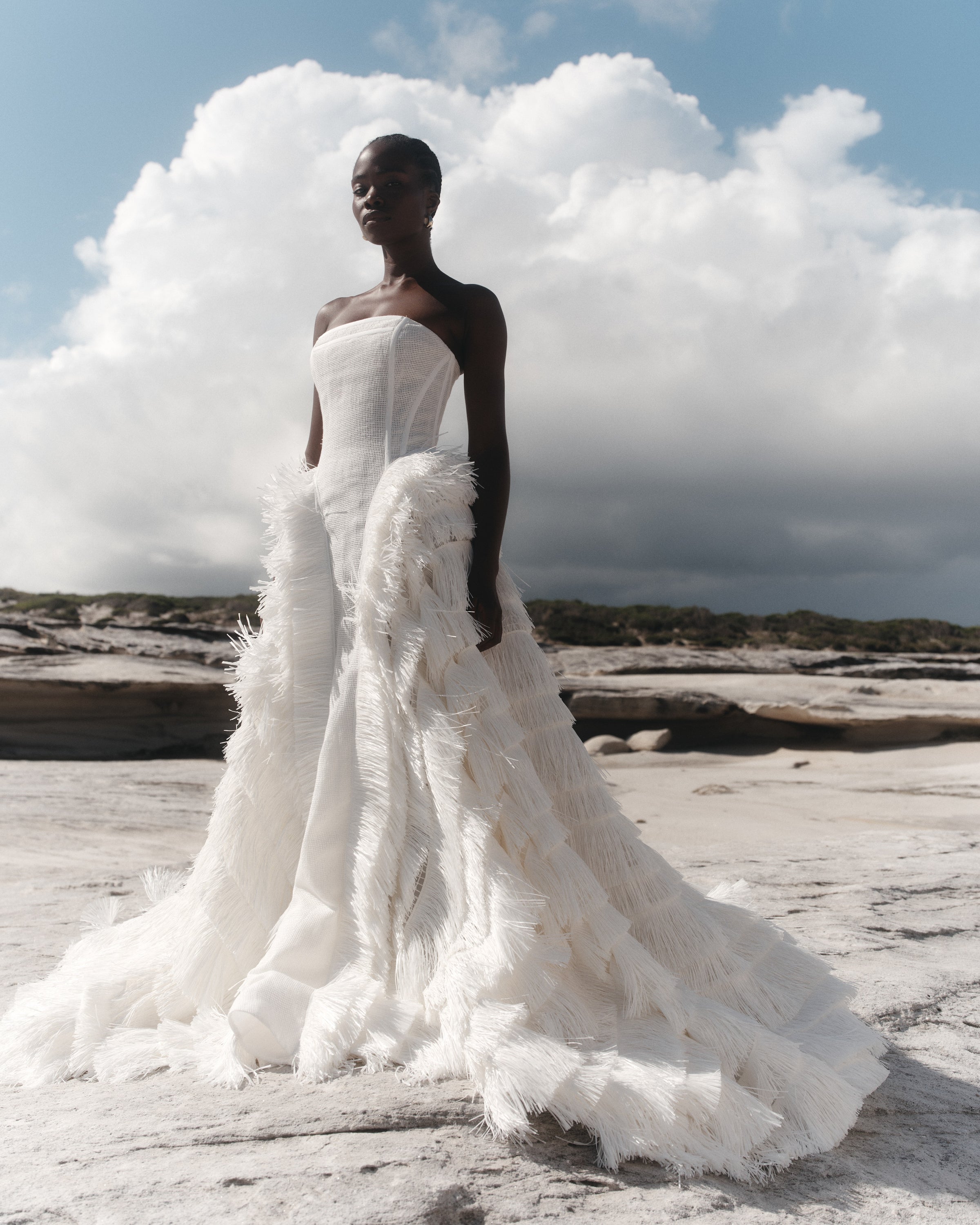 Woman in a white wedding dress with feathered details standing on a rocky beach.