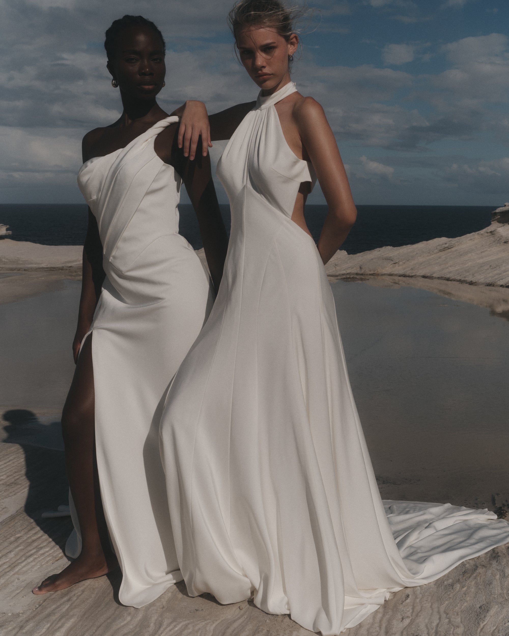 Two women in white dresses standing on a coastal cliff with ocean view.
